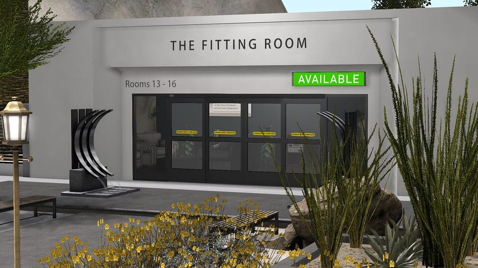 Exterior facade of The Fitting Room building. Clean rendered white walls, floor-to-ceiling dark glass automatic doors, signage reading Rooms 13–16, a green Available sign, desert landscaping with yellow flowering shrubs, agave, tall ornamental grasses, a brass lantern post, a curved black metal sculpture, and a bench seat along the building front.